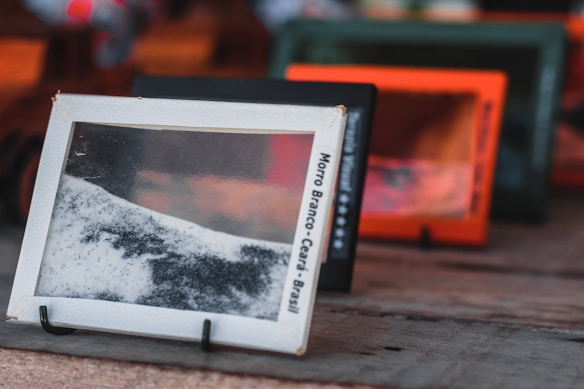 A series of rectangular frames made of what appears to be sand or fine particles encased in transparent covers are placed in a row, each sitting on small stands. The frames vary in color, with an emphasis on black, white, and shades of orange. They are positioned on a wooden surface, with the main frame prominently displaying a mix of black and white sand. The text on the visible frame reads 'Morro Branco - Cear&aacute; - Brasil.' The focus is on the foreground frame, with others receding into the background, creating a sense of depth.