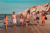 A group of six young people walks along a sunny beach, with several wearing swimwear and holding shoes in their hands. They are engaged in conversation and appear happy and relaxed. The beach has cliffs in the background and a few parked vehicles along the shoreline.