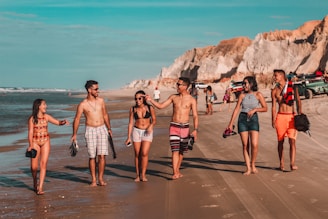 A group of six young people walks along a sunny beach, with several wearing swimwear and holding shoes in their hands. They are engaged in conversation and appear happy and relaxed. The beach has cliffs in the background and a few parked vehicles along the shoreline.
