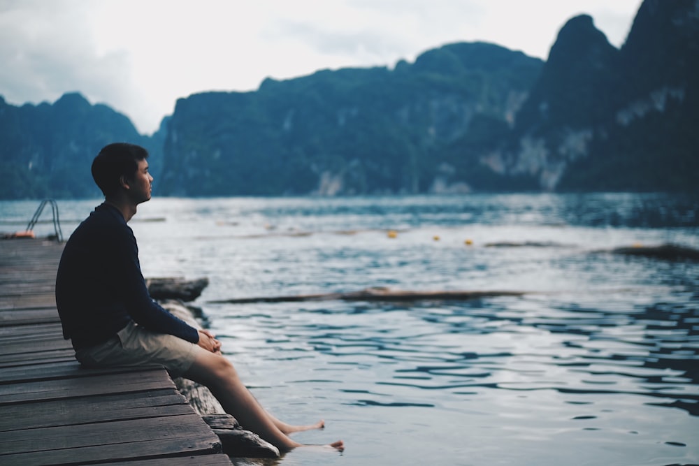 man in black jacket sitting on wooden dock during daytime