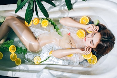 A woman relaxes in a bathtub filled with water, surrounded by floating slices of citrus fruits such as oranges and limes. Green fern leaves are also present in the tub, adding to the serene atmosphere. The woman has long hair and appears calm and peaceful.