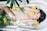 A woman relaxes in a bathtub filled with water, surrounded by floating slices of citrus fruits such as oranges and limes. Green fern leaves are also present in the tub, adding to the serene atmosphere. The woman has long hair and appears calm and peaceful.