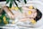 A woman relaxes in a bathtub filled with water, surrounded by floating slices of citrus fruits such as oranges and limes. Green fern leaves are also present in the tub, adding to the serene atmosphere. The woman has long hair and appears calm and peaceful.