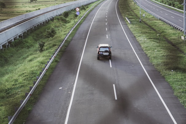 A solitary SUV travels along a wide, empty highway surrounded by green grass and trees. The road is well-maintained with clear white lane markings. Traffic signs are visible on the roadside, and there's a smooth curve in the road.