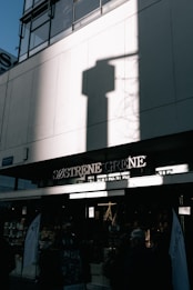The photo captures an urban storefront with the sign 'Søstrene Grene' under a tall building. A large shadow of a shape is cast on the building's white facade. The lower part of the image shows the entrance to the store with people entering or passing by.