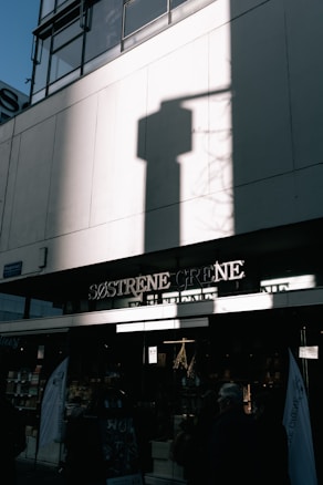 The photo captures an urban storefront with the sign 'Søstrene Grene' under a tall building. A large shadow of a shape is cast on the building's white facade. The lower part of the image shows the entrance to the store with people entering or passing by.