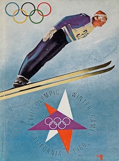A dynamic illustration of a ski jumper in mid-air, wearing a number 30 jersey, depicted against a blue backdrop. The Olympic rings are displayed prominently above, and the text ‘VIII Olympic Winter Games California 1960’ surrounds a star design at the bottom. Sketches of various winter sports are lightly integrated into the background.