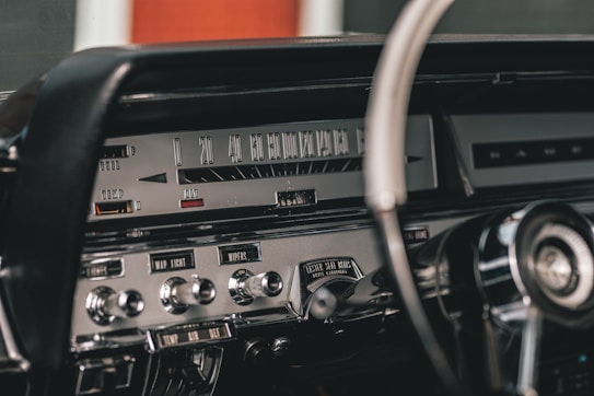 The image depicts the interior of a vintage car dashboard. The central focus is on the speedometer, which displays numerical speed values. There are various control knobs and switches labeled with functions such as 'MAP LIGHT', 'WIPERS', and movements related to 'WASHER' and 'LIGHTS'. A retro steering wheel is partially visible on the right side of the image. The dashboard and controls have a chrome and metallic finish, giving it a classic and polished look.