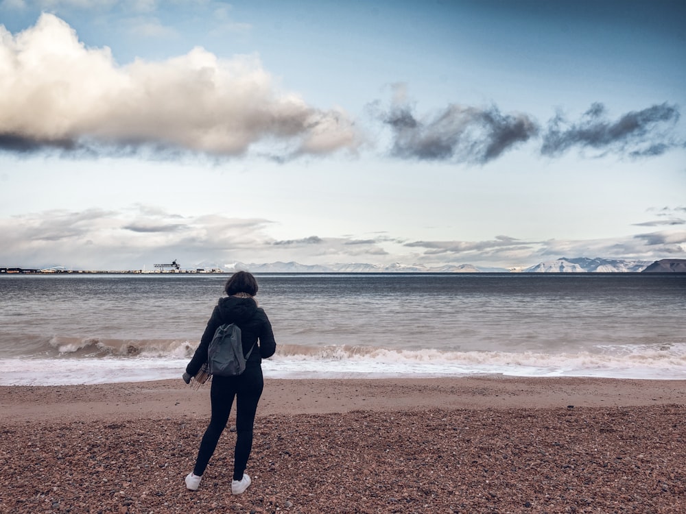 woman in black jacket and black pants standing on seashore during daytime