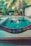 A tranquil backyard setting featuring a small, rectangular swimming pool surrounded by tropical foliage. A multicolored hammock is prominently displayed in the foreground. Behind the pool, there are lush green plants and trees, with laundry hanging on a line.