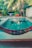 A tranquil backyard setting featuring a small, rectangular swimming pool surrounded by tropical foliage. A multicolored hammock is prominently displayed in the foreground. Behind the pool, there are lush green plants and trees, with laundry hanging on a line.
