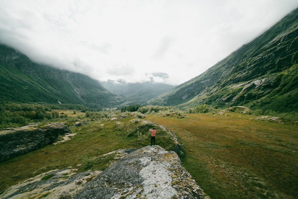 person in red jacket standing on gray concrete pathway near green mountains during daytime