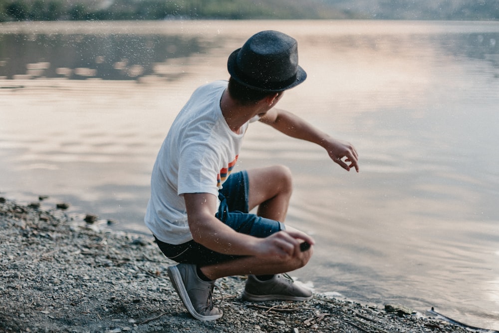 man in white t-shirt and blue denim shorts sitting on rock near body of water