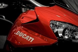 A close-up view of the front portion of a red Ducati motorcycle, highlighting its sleek design and the illuminated headlight. The brand name Ducati is prominently displayed on the red bodywork, conveying a sense of speed and style.