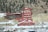 A large industrial building, labeled 'Argo Gold Mine & Mill,' stands prominently with its distinct red color against the backdrop of a rocky and forested hillside. The snowy landscape surrounds the structure, and a nearby commercial area includes a Subway restaurant. Trees dot the hills in the background.