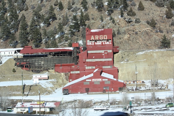 A large industrial building, labeled 'Argo Gold Mine & Mill,' stands prominently with its distinct red color against the backdrop of a rocky and forested hillside. The snowy landscape surrounds the structure, and a nearby commercial area includes a Subway restaurant. Trees dot the hills in the background.