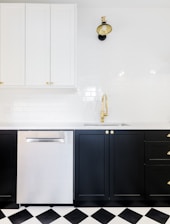 A modern kitchen interior featuring black lower cabinets with gold handles, a stainless steel dishwasher, and white upper cabinets. The backsplash is made of white subway tiles. A stylish black and gold wall lamp is mounted above a gold faucet, and the floor has a black and white checkered pattern.