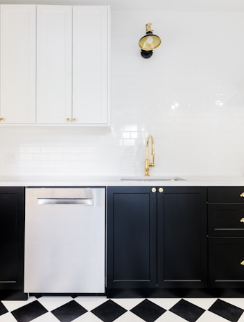 A modern kitchen interior featuring black lower cabinets with gold handles, a stainless steel dishwasher, and white upper cabinets. The backsplash is made of white subway tiles. A stylish black and gold wall lamp is mounted above a gold faucet, and the floor has a black and white checkered pattern.