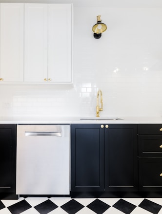 A modern kitchen interior featuring black lower cabinets with gold handles, a stainless steel dishwasher, and white upper cabinets. The backsplash is made of white subway tiles. A stylish black and gold wall lamp is mounted above a gold faucet, and the floor has a black and white checkered pattern.