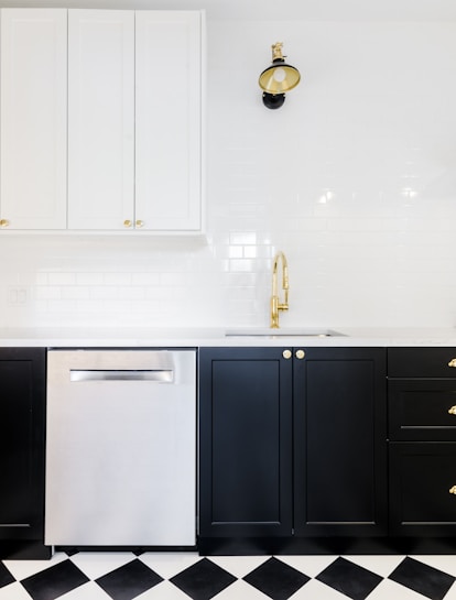 A modern kitchen interior featuring black lower cabinets with gold handles, a stainless steel dishwasher, and white upper cabinets. The backsplash is made of white subway tiles. A stylish black and gold wall lamp is mounted above a gold faucet, and the floor has a black and white checkered pattern.