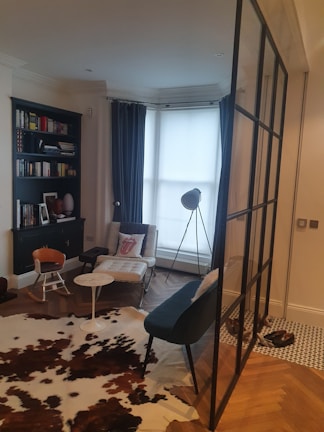 A cozy, modern living space features a large window with soft blue curtains, a black bookshelf filled with books, and a white lounge chair with a decorative pillow. A stylish tripod floor lamp stands near the window, illuminating the area. The floor is a mix of wooden herringbone and patterned tiles, with an accent cowhide rug. A small round table separates an orange and an elegant gray chair. A glass partition adds a contemporary touch to the room.