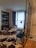 A cozy, modern living space features a large window with soft blue curtains, a black bookshelf filled with books, and a white lounge chair with a decorative pillow. A stylish tripod floor lamp stands near the window, illuminating the area. The floor is a mix of wooden herringbone and patterned tiles, with an accent cowhide rug. A small round table separates an orange and an elegant gray chair. A glass partition adds a contemporary touch to the room.
