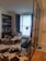 A cozy, modern living space features a large window with soft blue curtains, a black bookshelf filled with books, and a white lounge chair with a decorative pillow. A stylish tripod floor lamp stands near the window, illuminating the area. The floor is a mix of wooden herringbone and patterned tiles, with an accent cowhide rug. A small round table separates an orange and an elegant gray chair. A glass partition adds a contemporary touch to the room.