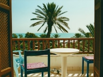 A balcony with wicker doors opens to a scenic view of the ocean. A palm tree stands prominently in the center, surrounded by other lush greenery. The foreground has a round white table and two blue chairs with patterned cushions. A pair of shoes rests on the floor near the table.
