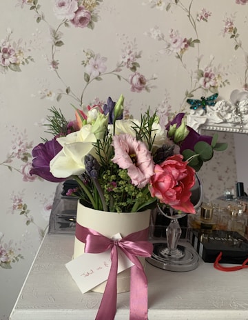 A decorative bouquet of flowers is arranged in a cream-colored cylindrical vase adorned with a pink ribbon. The bouquet contains a variety of flowers, including white, pink, purple, and red blossoms, with accents of greenery. A small card is attached to the vase with handwriting visible. The background features a floral-patterned wallpaper with pastel tones. A table holds various items, including a mirror and beauty products.