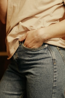 A close-up view of a person wearing a beige shirt tucked into blue denim jeans, with their hand slipped casually into the front pocket. The fabric of the jeans shows classic blue denim texture.