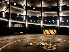 A multi-level parking garage with fluorescent lights illuminating the structure. The image features several floors with metal railings, and a concrete surface marked with a bicycle symbol. A yellow and black striped barrier is visible on the ground.