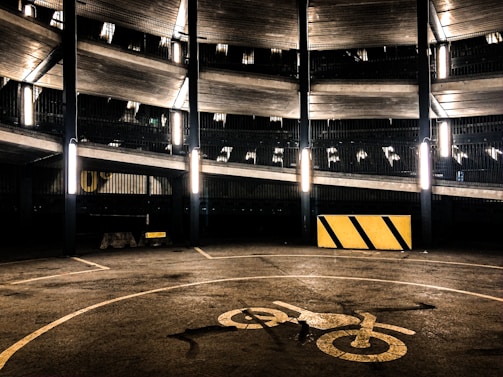 A multi-level parking garage with fluorescent lights illuminating the structure. The image features several floors with metal railings, and a concrete surface marked with a bicycle symbol. A yellow and black striped barrier is visible on the ground.
