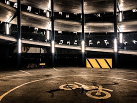 A multi-level parking garage with fluorescent lights illuminating the structure. The image features several floors with metal railings, and a concrete surface marked with a bicycle symbol. A yellow and black striped barrier is visible on the ground.