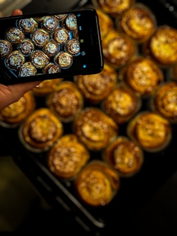 A hand is holding a smartphone taking a photo of several freshly baked pastries arranged neatly. The pastries have a golden-brown color and are topped with a sprinkling of what appears to be sugar or seeds. The phone's screen displays the pastries.