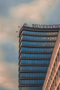 A modern skyscraper with a series of reflective blue-tinted glass windows curves elegantly against an overcast sky. The top of the building features a sign with the partially visible word 'SILVERTOWER'. The structure's facade displays a seamless and sleek architectural design, conveying a sense of sophistication and contemporary style.