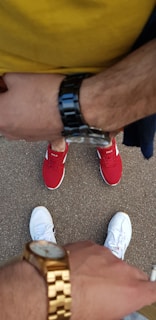 Two individuals are standing on a textured pavement, each wearing distinct watches and sneakers. The top individual's arm features a dark banded watch, and they are wearing red sneakers with a 'POLO' logo. The lower individual displays a gold watch and is wearing white sneakers.