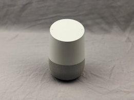 A minimalist smart speaker with a cylindrical design featuring a matte white top and a grey fabric-covered base, set against a plain background.