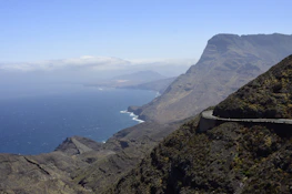 A coastal mountain road with a van winding through, a solitary male figure standing nearby taking in the view.