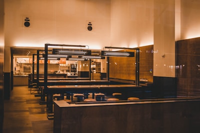 A modern, minimalist restaurant interior features wooden tables and stools, with soft, warm lighting creating an inviting ambience. The space includes a counter with overhead lighting and visible menu boards, suggesting a fast-casual dining environment.