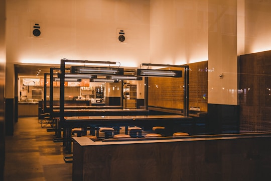 A modern, minimalist restaurant interior features wooden tables and stools, with soft, warm lighting creating an inviting ambience. The space includes a counter with overhead lighting and visible menu boards, suggesting a fast-casual dining environment.