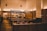 A modern, minimalist restaurant interior features wooden tables and stools, with soft, warm lighting creating an inviting ambience. The space includes a counter with overhead lighting and visible menu boards, suggesting a fast-casual dining environment.