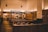 A modern, minimalist restaurant interior features wooden tables and stools, with soft, warm lighting creating an inviting ambience. The space includes a counter with overhead lighting and visible menu boards, suggesting a fast-casual dining environment.