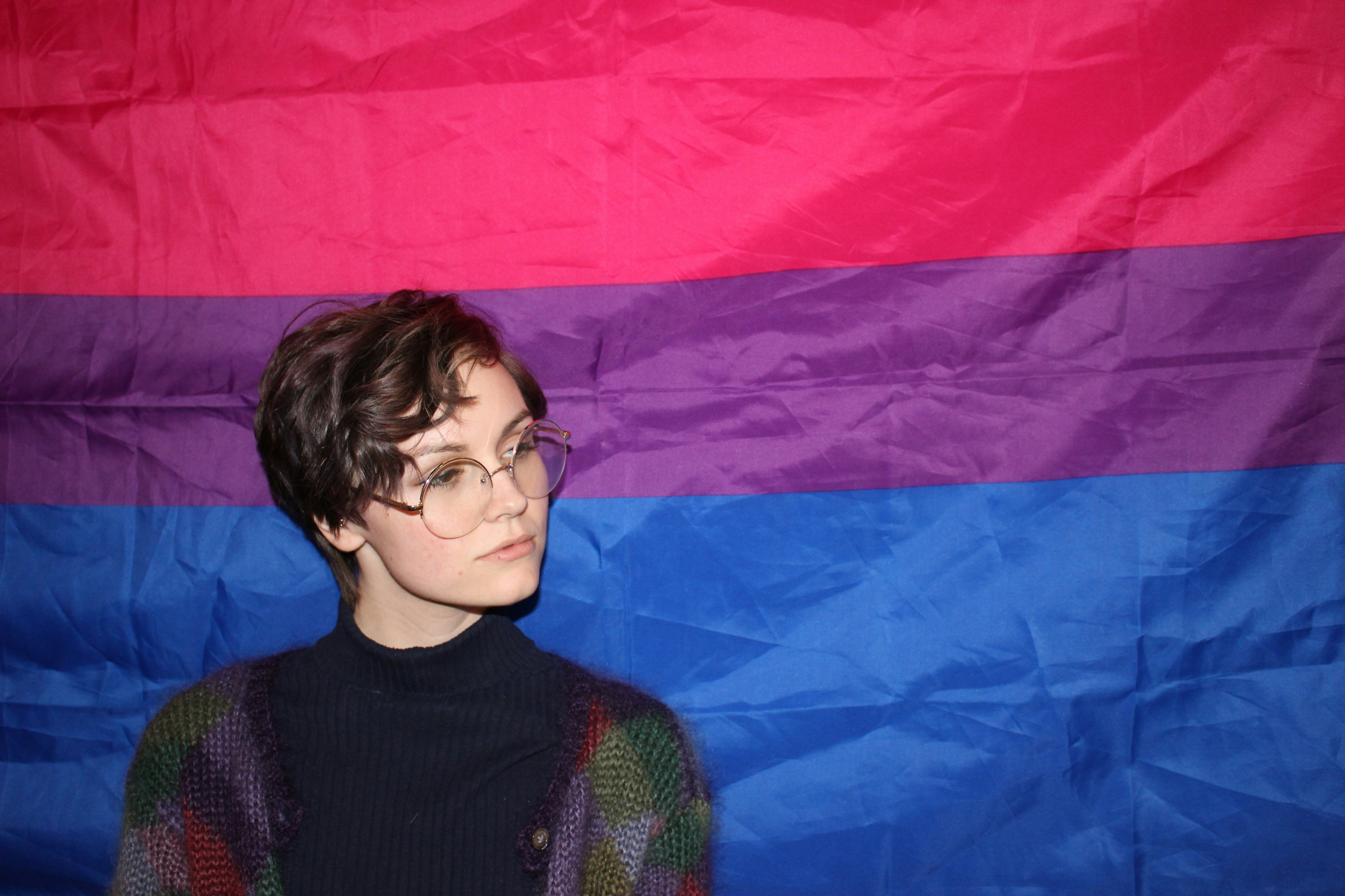 A femme-presenting individual with wire-rimmed glasses looks thoughtfully off-camera in front of a bisexual pride flag.