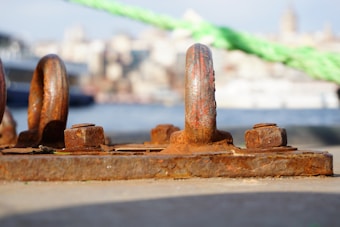A close-up view of a rusty metal mooring ring and bolts on a dock, with an out-of-focus view of water and a cityscape in the background. The metal is heavily corroded, showing signs of wear, while a green rope is visible across the scene.
