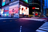 A cityscape scene at night with bright, colorful advertising billboards lighting up the streets. The large ads include brands like Walgreens, Westworld, and H&M, with an illuminated street below and crosswalks leading across an empty intersection.