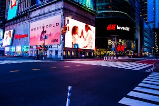 A cityscape scene at night with bright, colorful advertising billboards lighting up the streets. The large ads include brands like Walgreens, Westworld, and H&M, with an illuminated street below and crosswalks leading across an empty intersection.