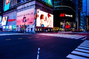 A cityscape scene at night with bright, colorful advertising billboards lighting up the streets. The large ads include brands like Walgreens, Westworld, and H&M, with an illuminated street below and crosswalks leading across an empty intersection.