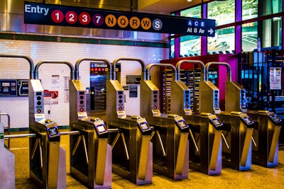 Turnstiles at a subway station are positioned in a row, with electronic card readers on top for entry. Above, a large sign displays various subway lines indicated by colored circles with numbers and letters. The surroundings are lit with artificial lighting, and the walls are tiled in white.