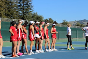 A group of women in red athletic uniforms and white visors stand in a line on a tennis court. They appear to be listening to a person dressed in casual athletic wear, who is standing to the right of the group. The background includes a green fence and trees, as well as distant hills under a clear blue sky.