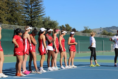 A group of women in red athletic uniforms and white visors stand in a line on a tennis court. They appear to be listening to a person dressed in casual athletic wear, who is standing to the right of the group. The background includes a green fence and trees, as well as distant hills under a clear blue sky.